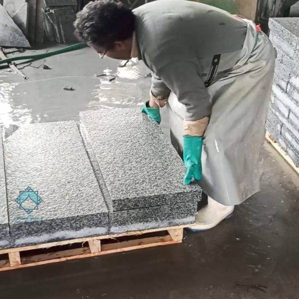 کارگر در حال بررسی و مرتب‌سازی جدول سنگی و سنگ حجمی در کارخانه تولید سنگ، در کنار پالت‌های چوبی پر از سنگ‌های گرانیت