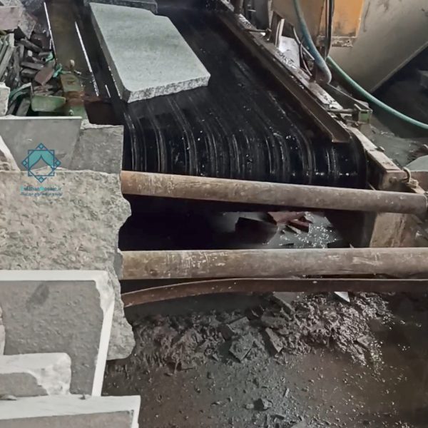 نوار نقاله در حال انتقال جدول‌های سنگی و سنگ‌های حجمی در کارخانه تولید سنگ، با سنگ‌های گرانیت و محیط صنعتی پر از ابزارآلات